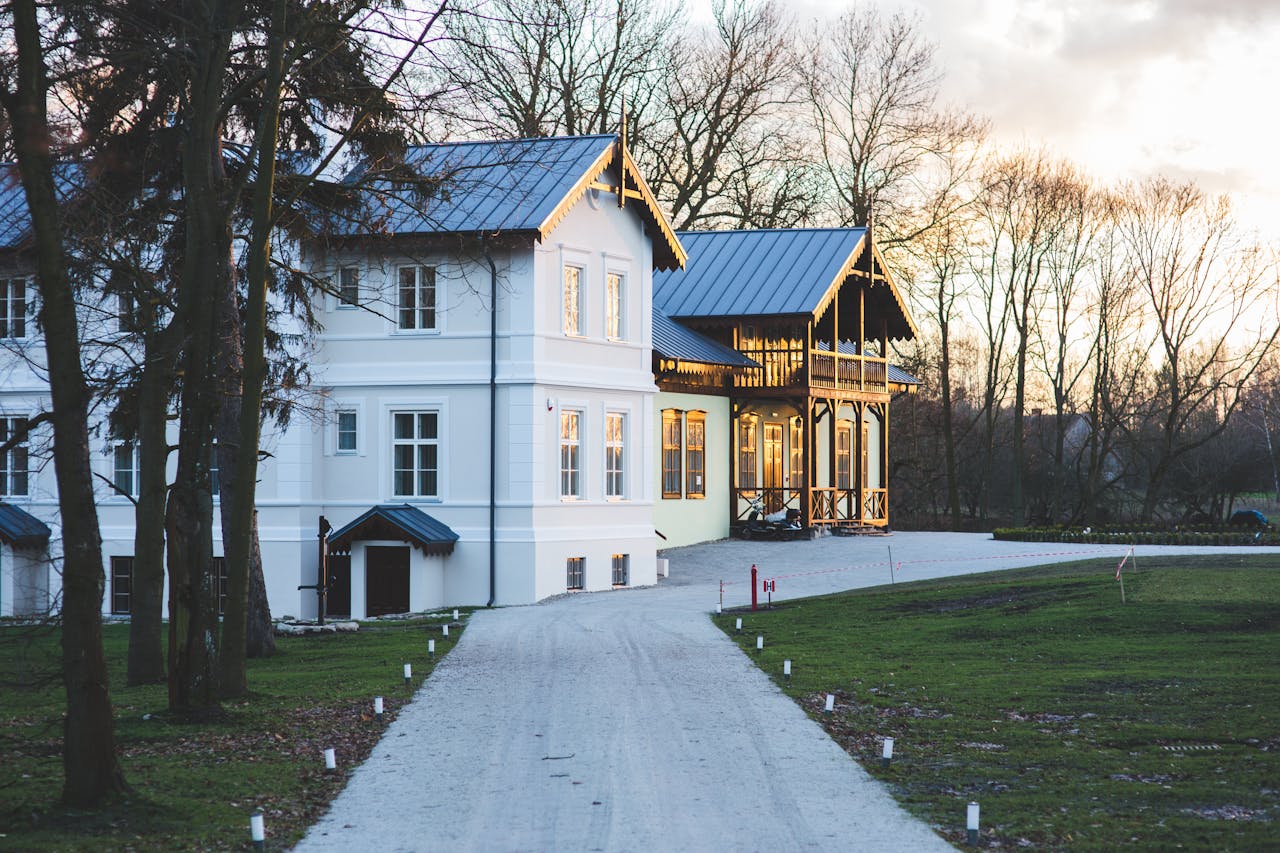 White mansion with a garden and driveway at sunset, showcasing elegant architecture.